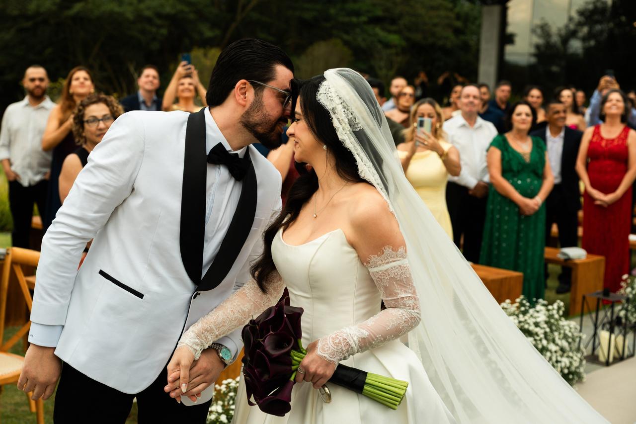 Noivo de smoking branco com lapela preta e noiva de véu, de testas encostadas no altar, diante dos convidados