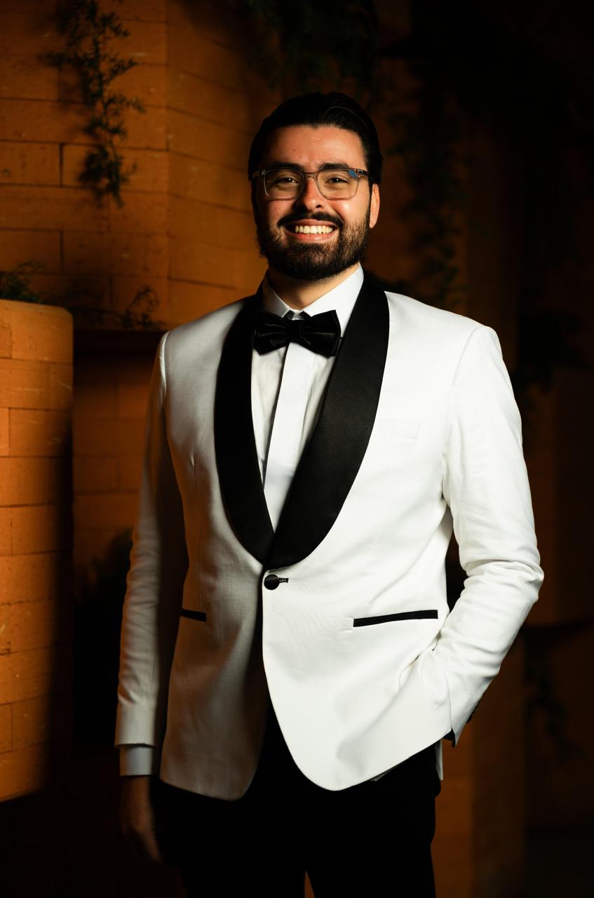 Noivo de smoking branco com lapela xale preta e gravata borboleta, sorrindo, com a mão no bolso