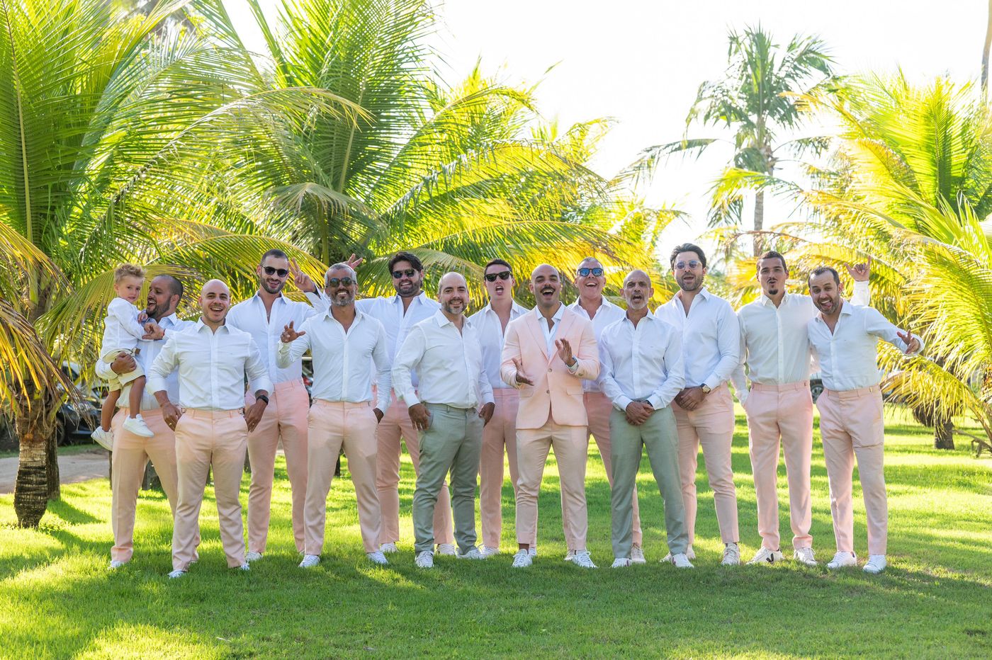 Noivo de blazer rosa-claro entre os padrinhos, todos de camisa branca e calça rosa ou verde-acinzentada, em um coqueiral