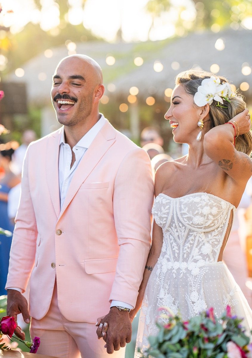Noivo de terno rosa-claro rindo, de mãos dadas com a noiva de vestido de renda e flor branca no cabelo
