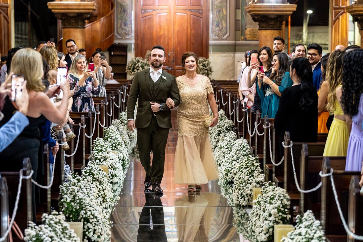Noivo de terno verde-musgo entrando pelo corredor da igreja, ladeado de flores brancas, com uma senhora de vestido dourado