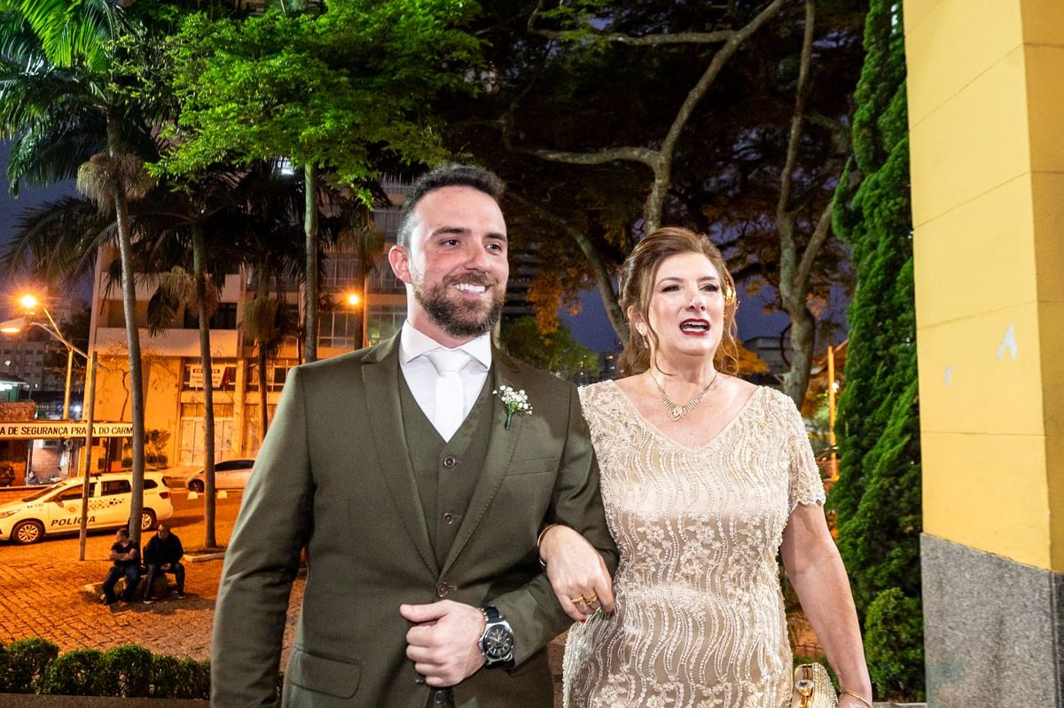 Noivo de terno verde-musgo sorrindo, de braço dado com uma senhora de vestido dourado, à noite do lado de fora da igreja