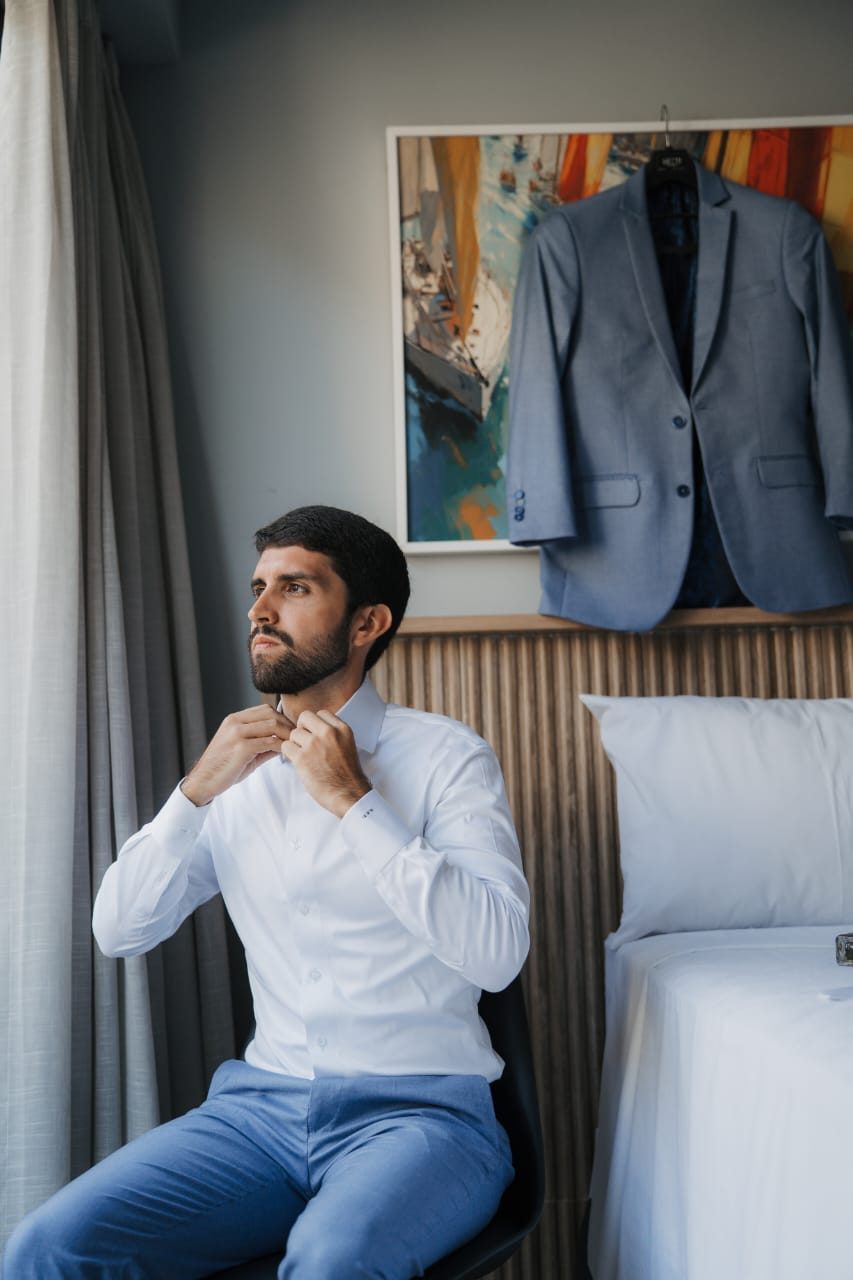 Noivo sentado ajustando o colarinho da camisa branca, com o paletó azul-acinzentado pendurado ao fundo