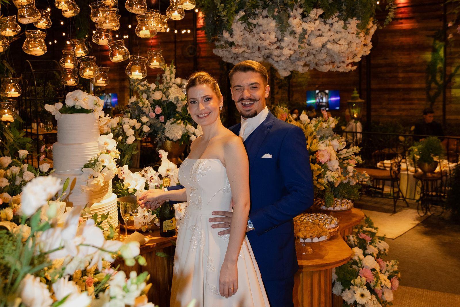 Noivo de terno azul-marinho de três peças ao lado da noiva, cortando o bolo do casamento sob velas suspensas e flores brancas