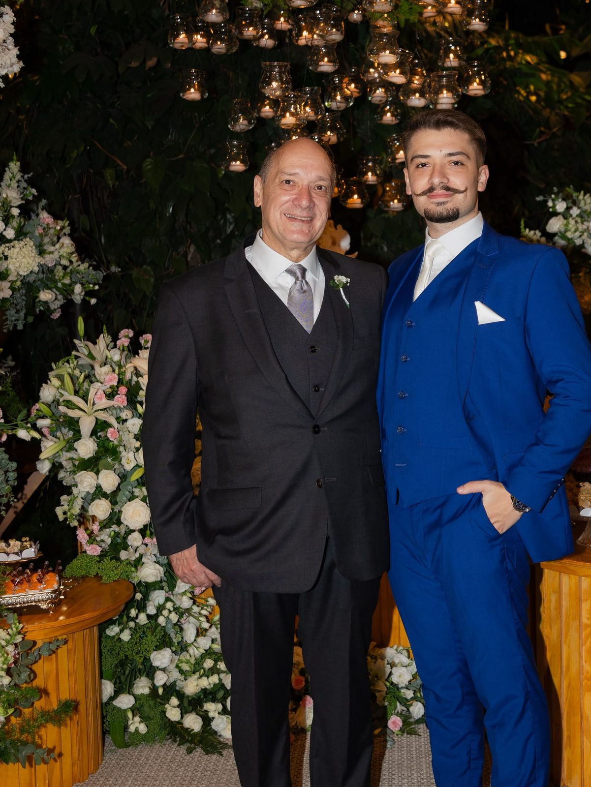 Adriano, de terno azul royal de três peças, ao lado do pai, de terno cinza-chumbo, sob um arranjo de flores brancas e velas