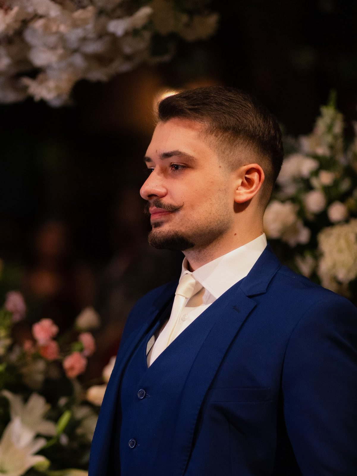Noivo de terno azul royal de três peças e gravata off-white, em perfil, diante de um arranjo de flores brancas