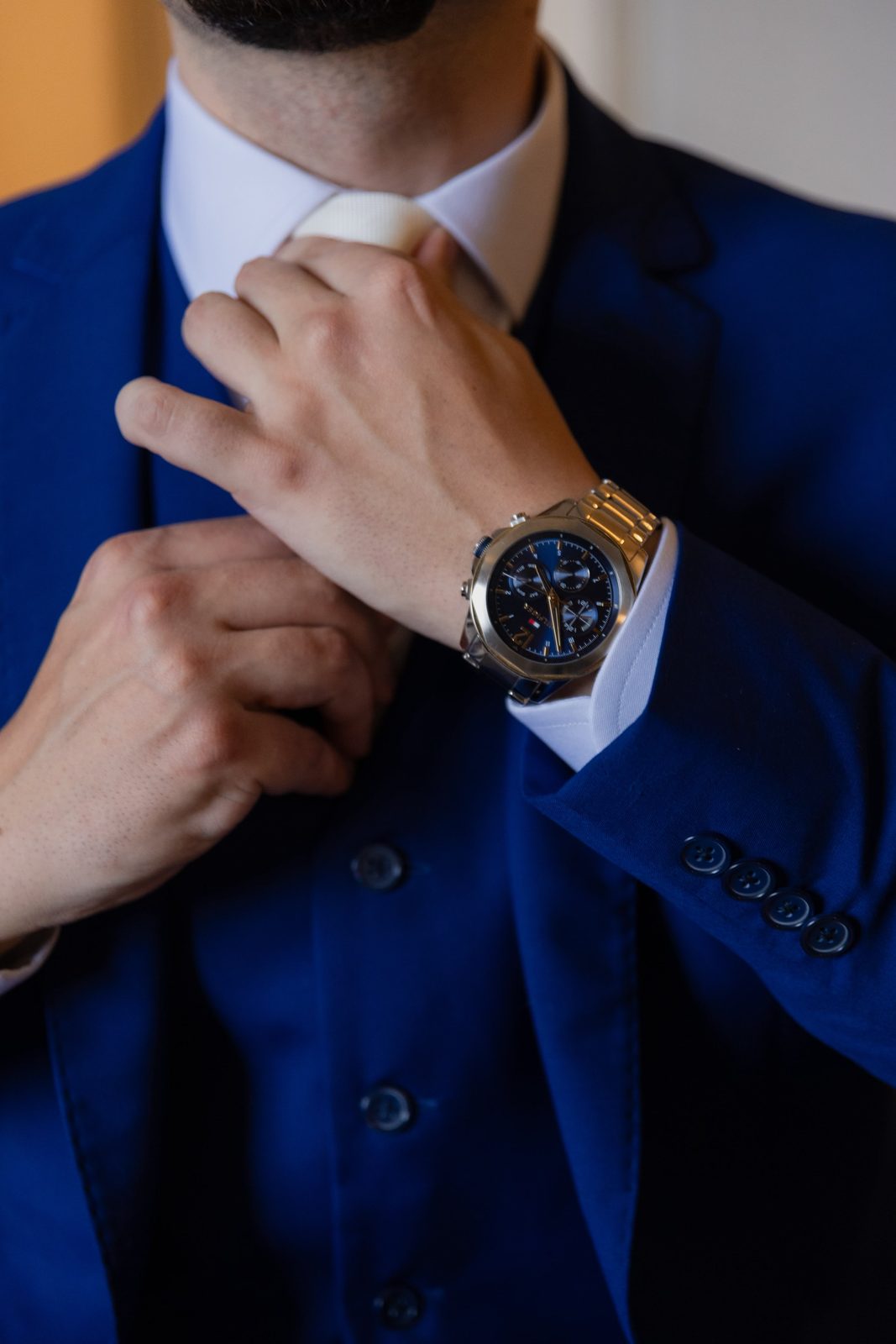 Detalhe das mãos ajustando a gravata, com relógio dourado no punho e terno azul royal