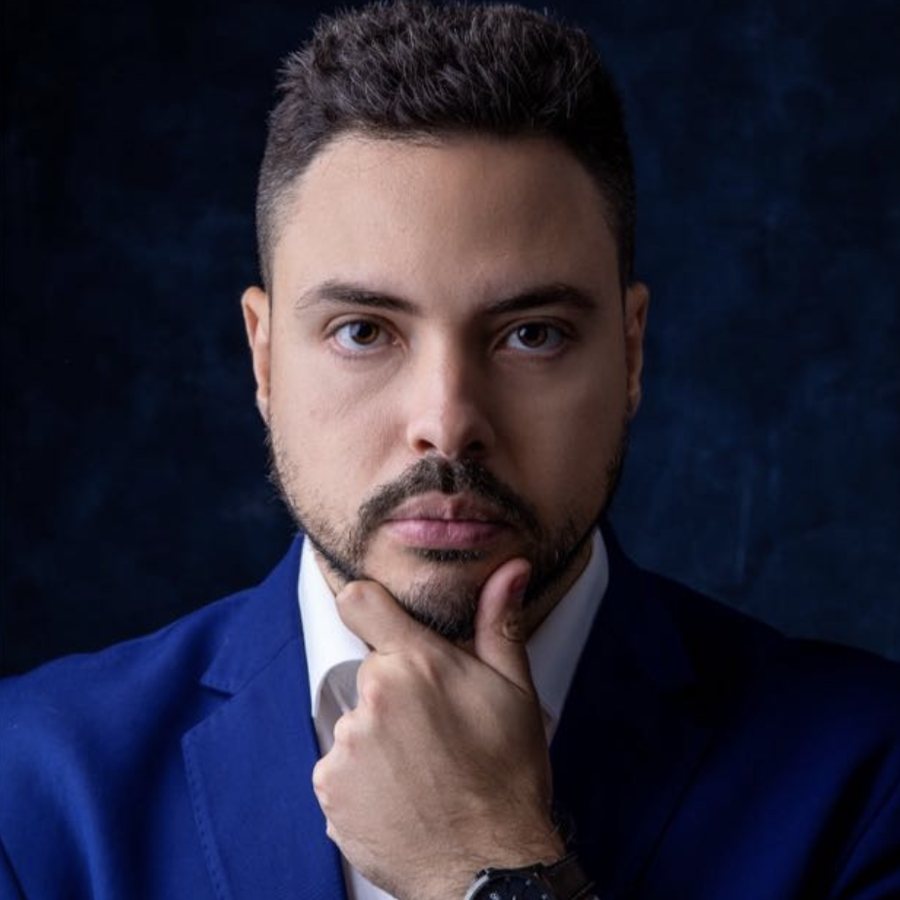 Retrato de Danilo Noronha, de terno azul e mão no queixo, em fundo escuro